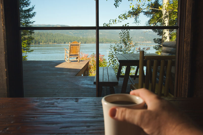 A lake viewed through a picture window in a lake home by a man drinking coffee.