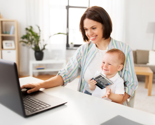 A woman sitting at a computer in a home office, holding a baby.