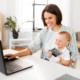 A woman sitting at a computer in a home office, holding a baby.