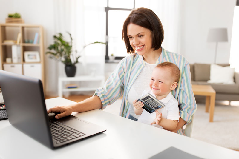 A woman sitting at a computer in a home office, holding a baby.